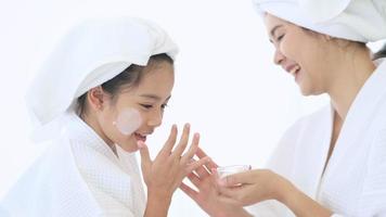 Happy beautiful mom and daughter in white bathrobe applying moisturizing cream on face at home, skin care and treatment concept video