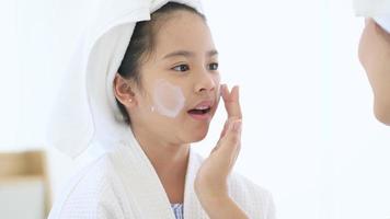 Happy beautiful mom and daughter in white bathrobe applying moisturizing cream on face at home, skin care and treatment concept video