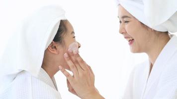 Happy beautiful mom and daughter in white bathrobe applying moisturizing cream on face at home, skin care and treatment concept video