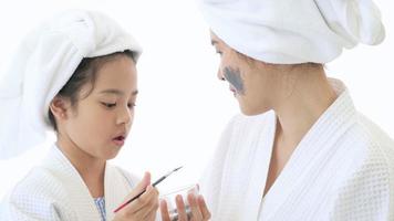 Happy beautiful mom and daughter in white bathrobe applying face mask in bedroom, Family and Beauty Concept.
