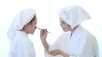 Happy beautiful mom and daughter in white bathrobe applying face mask in bedroom, Family and Beauty Concept. video