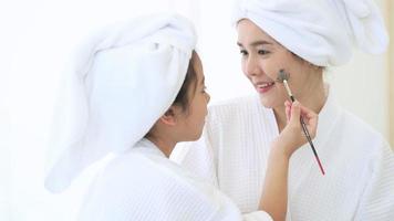 Happy beautiful mom and daughter in white bathrobe applying face mask in bedroom, Family and Beauty Concept.