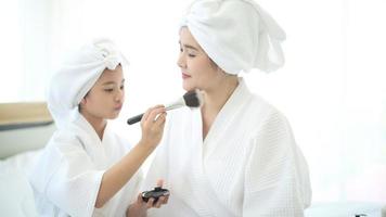 feliz linda mãe e filha em roupão branco aplicando maquiagem natural com pincel de pó cosmético, família e conceito de beleza.