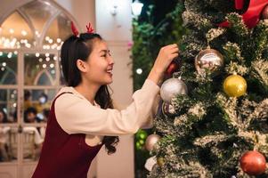 Asian woman wearing antler headband decorating ornament ball on christmas tree