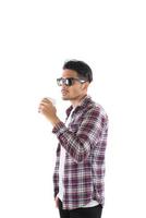 retrato de un apuesto joven hipster de pie, con gafas de sol, camisa y sosteniendo una taza de café sobre un fondo blanco aislado. foto