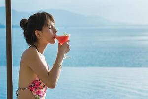 Beautiful young women in bikini lying on a deckchair pool with a red drink by the sea photo