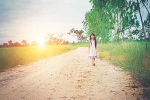 una niña pequeña con el pelo largo vestido se aleja de ti por un camino rural.