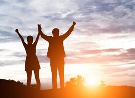 Silhouette of young couple together arms up in the air of happiness with accomplishment in the clouds at sunset.
