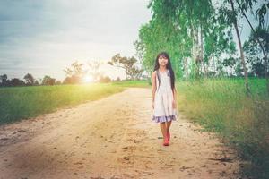 Little girl with long hair wearing dress is walking away from you down rural road. photo