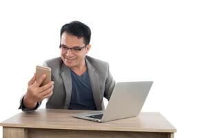 Confident young man working on laptop and looking on the mobile phone while sitting isolated on white background. photo