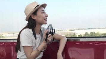 Beautiful Asian female tourist with camera sits in a red seat, traveling by train, taking snapshot photo when transporting in suburb view, enjoy passenger lifestyle by railway, happy journey vacation. video