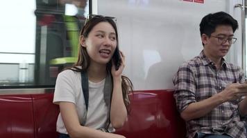 vacker asiatisk kvinnlig turist med kamera sitter i rött säte, reser med skytrain, pratar mobiltelefon när du transporterar i stadsvy, stadspassagerarlivsstil med järnväg, lycklig resa semester. video