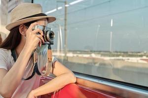 Beautiful Asian female tourist with face mask sits in a red seat, traveling by train, taking snapshot photo, transporting in suburb view, enjoy passenger lifestyle by railway, happy journey vacation. photo