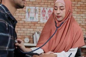 Young beautiful woman doctor is health examining male patient in office of hospital clinic and advising with a smile on medicines. This Asian medical specialist is an Islamic person wearing a hijab. photo
