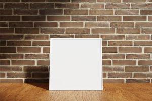 Square white poster or photo frame mockup on the wooden table with blurry brick background