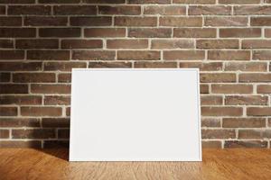 Horizontal white poster or photo frame mockup on the wooden table with blurry brick background