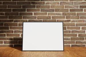 Horizontal black poster or photo frame mockup on the wooden table with blurry brick background