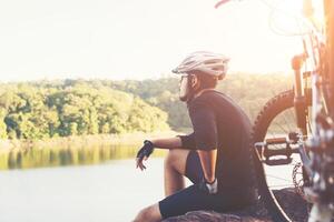 Young athlete man sitting on a rock with bicycle enjoy with nature of lake during sunset. photo