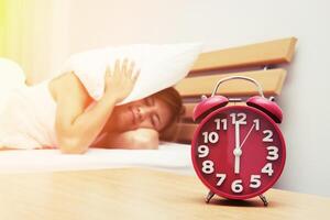 Young beautiful woman hates waking up early in the morning. Sleepy girl looking at alarm clock and trying to hide under the pillow in the morning photo