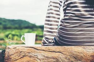 Back of young woman with white of hot coffee cup sitting on the bench with green nature mountain background. photo