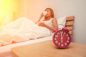 Young beautiful woman hates waking up early in the morning. Sleepy girl looking at alarm clock and trying to hide under the pillow in the morning photo