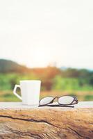 White of hot coffee cup with glasses on the bench with green nature mountain background. photo