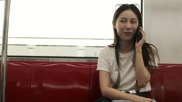 linda turista asiática com câmera senta-se no assento vermelho, viajando de trem do céu, falando no celular ao transportar em vista urbana, estilo de vida de passageiros da cidade por via férrea, férias de viagem feliz. video