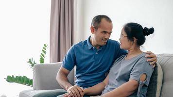 Portrait of Asian mature couple sitting in the living room. wife and husband hugging, holding hand with happiness and cheerful. Love, safety, and insurance family concept photo