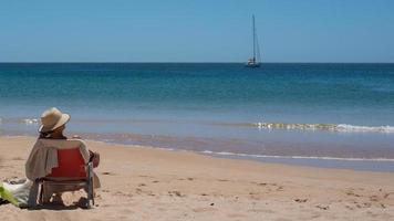 Woman sitting on a chair with a hat, on the beach and looking to the horizon. Photo taken from her back. A yacht in the distance. Portugal