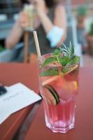 agua de pepino con bayas y rodaja de limón, con color rosa y una pajita de cartón. mujer en el fondo con otro vaso. foto