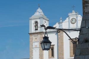 Close up of a street lamp with a dragon sculpture. In the background church at Giraldo square, Evora, Portugal. Blue sky photo