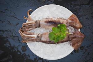 calamares frescos, pulpo o sepia para comida cocinada en el restaurante o en el mercado de mariscos, calamares crudos sobre hielo con ensalada de cilantro en el fondo de la placa blanca foto