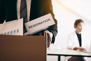 Front view with the male employee standing holding office supplies in the paper box going to submit a resignation letter while a female employee is working behind. photo