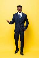 Full lenght portrait of young handsome African man in business suit smiling and opening his palm in yellow isolated studio background photo