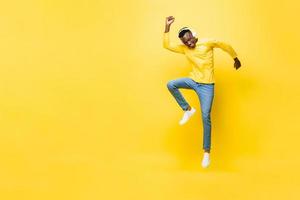 Happy energetic young African man wearing headphones listening to music and jumping with hand up next to empty space in yellow isolated studio background photo