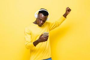 joven africano vestido de forma informal con auriculares inalámbricos escuchando música con un teléfono móvil en un estudio amarillo aislado foto