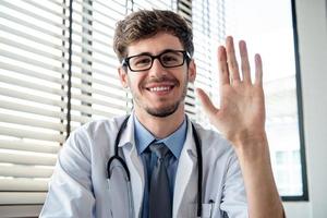 Smiling young male doctor waving hand to camera greeting patient online via distance video calling from the office photo