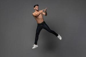 Handsome shirtless smiling Caucasian man jumping and pointing hands to copy space aside in gray isolated studio background photo