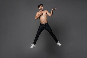 Handsome shirtless smiling Caucasian man jumping with hand opening to empty space aside in gray isolated studio background photo