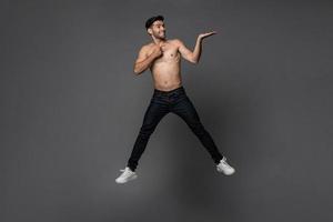 un apuesto hombre caucásico sonriente sin camisa saltando con la mano abriéndose a un espacio vacío a un lado en un estudio gris aislado foto