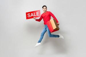Young Asian man holding shopping bags and red sale sign jumping in isolated light gray studio background for Chinese new year sale concept photo