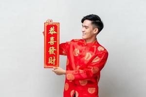 Handsome smiling Asian man showing red scroll  in isolated studio light gray background for Chinese new year concepts, foreign text means may you attain greater wealth photo