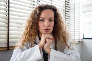 Serious female doctor carefully listening and keeping hands under chin while making medical conference video call from hospital, telehealth concept photo