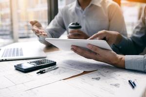 Two engineering working together and using digital tablet looking blueprint and analysis with architectural plan on desk. photo