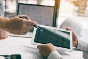 Two engineering working together and using digital tablet looking blueprint and analysis with architectural plan on desk. photo