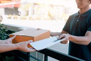 joven asiático sonriendo mientras entrega una caja de cartón a la mujer que tiene el documento para firmar la firma. foto