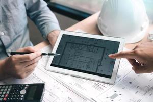 Two engineering working together and using digital tablet looking blueprint and analysis with architectural plan on desk. photo