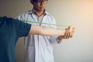 Ascendencia de fisioterapeuta masculino asiático trabajando y ayudando a proteger las manos de los pacientes con el paciente haciendo ejercicio de estiramiento con una banda de ejercicio flexible en la sala de la clínica. foto