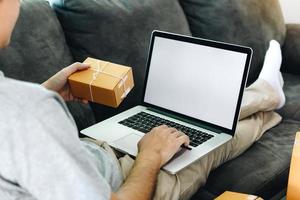 un hombre asiático sostiene una caja de entrega para el envío y está usando una pantalla en blanco de una computadora portátil. foto