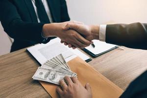 Two corporate businessmen shaking hands while one man places money on envelope document in office room with corruption concept. photo